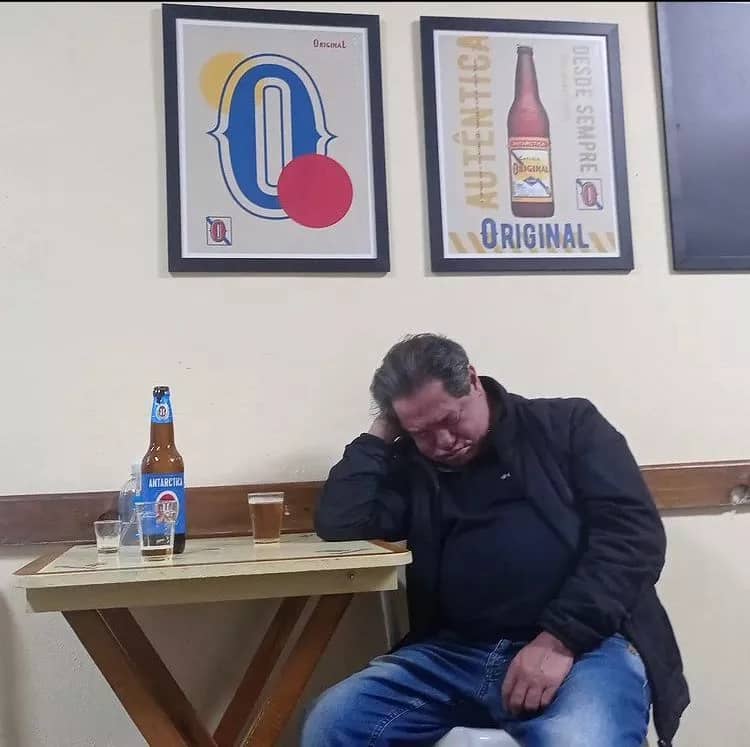 Foto do Pedrinho, dormindo sentado com a cabeça apoiada em uma das mãos, em uma mesa do Bar do Dionisio. Na mesa há uma garrafa de cerveja Antartica, dois copos vazios e um copo cheio. Pedrinho veste uma calça Jeans e uma blusa de nylon preta. Ao fundo, na parede, quadros da cerveja Original.