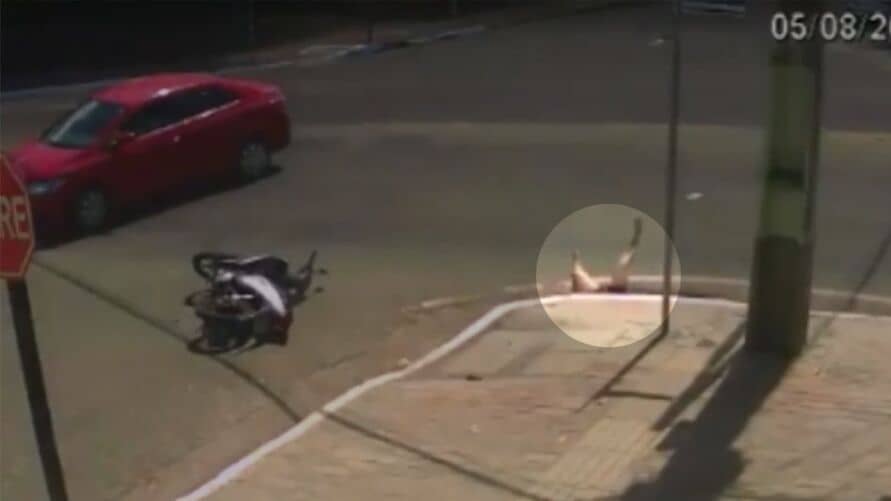 Foto do acidente onde uma mulher foi arremessada por um carro vermelho para dentro de um bueiro. Na imagem é possível ver o carro no canto superior direito, no meio do cruzamento, a moto caída no chão e em destaque a mulher caindo com as pernas para o alto no bueiro