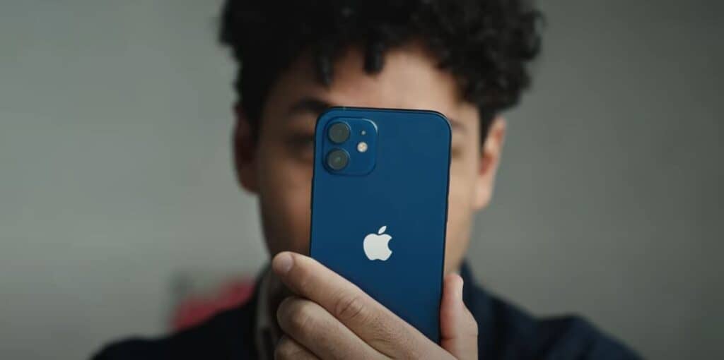 Foto de divulgação da campanha Privacidade no iPhone onde aparece um rapaz negro ao fundo desfocado segurando u iPhone 12 azul com uma das mãos.