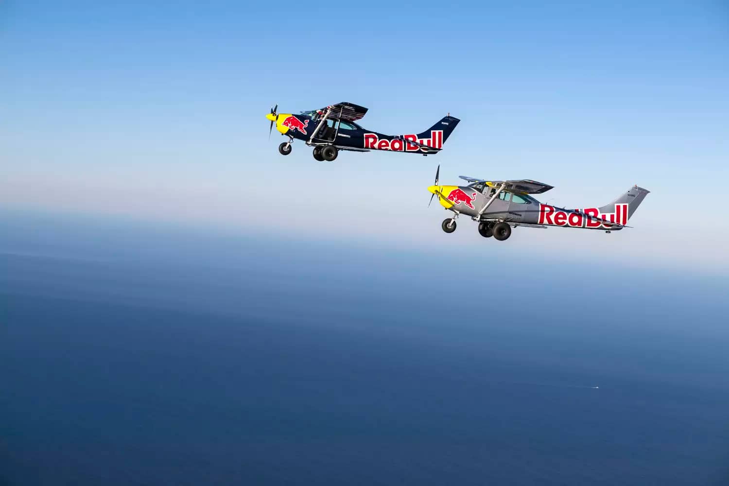 Foto dos dois aviões personalizados voando com a logo e brasão da marca Red Bull. Um avião é azul e o outro prateado, cores símbolo da companhia de energéticos.