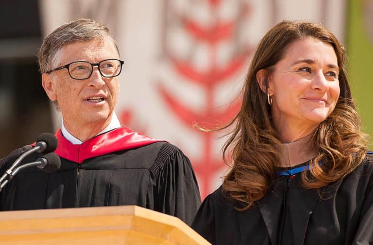 Bill e Melinda Gates, lado a lado vestidos de formandos em um púlpito em discurso de formatura. Ambos estão olhando para a direita para o horizonte.