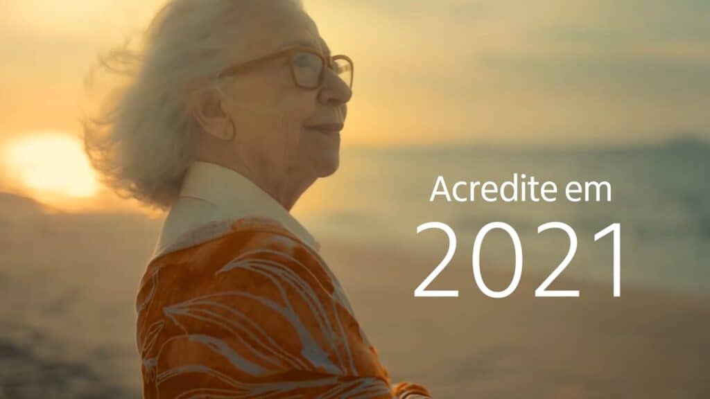 Imagem de Fernanda Montenegro onde aparece ao fundo o pôr do sol em uma praia deserta. Fernanda aparece de cabelos grisalhos, olhando para o horizonte e vestindo um casaco laranjado. Ao lado a legenda: Acredite em 2021 escrito em branco.