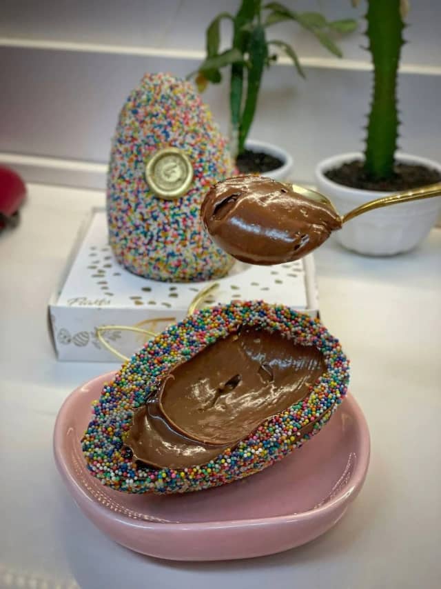 Na imagem aparece o ovo de Páscoa da Britney, com a casca cravejada com drageas colorias e recheado com brigadeiro gourmet. Uma colher dourada retirando o recheio do doce.